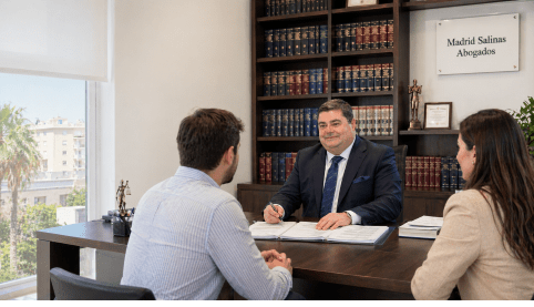 Abogado reunido con dos clientes en despacho jurídico con librería legal al fondo.